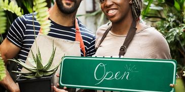 Two smiling people holding a green 'Open' sign and a potted plant.