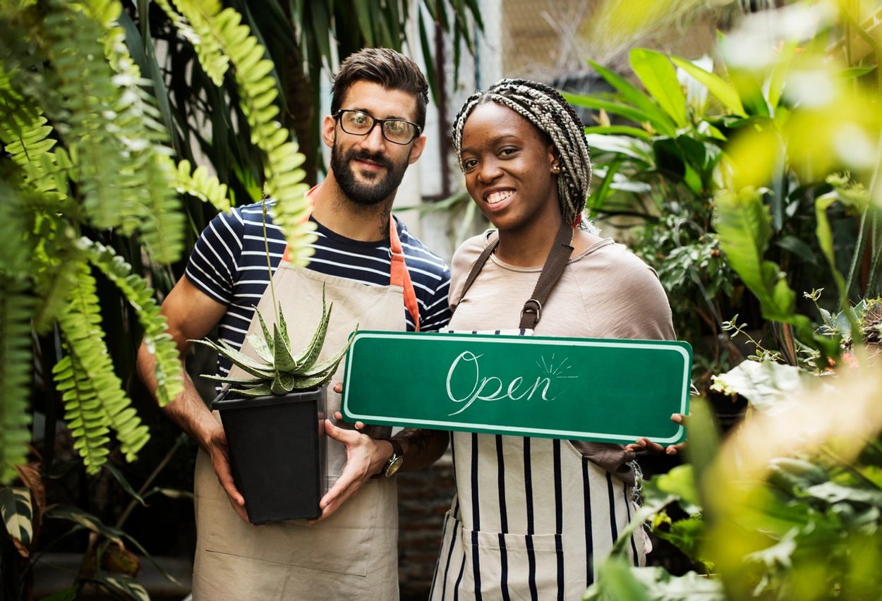 Two plant shop owners smiling and holding an "Open" sign and a potted plant.