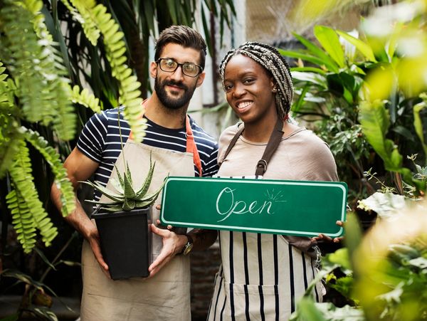 Two smiling shop owners hold a plant and an open sign in a lush garden store.