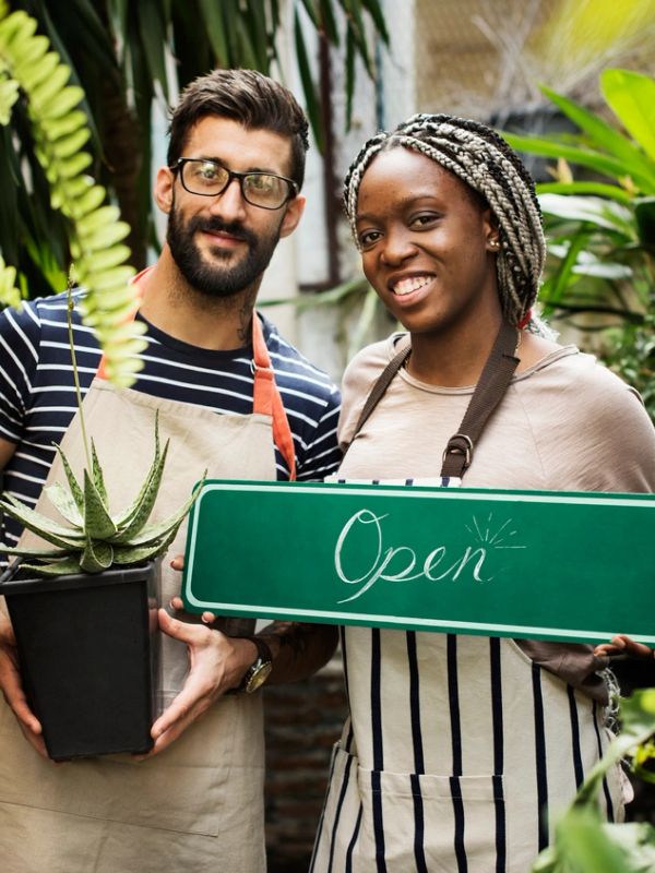 Two people smiling with open sign and plant.