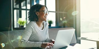 Woman with black hair smiling and seated with her hands on keyboard of a laptop, sunlight streams