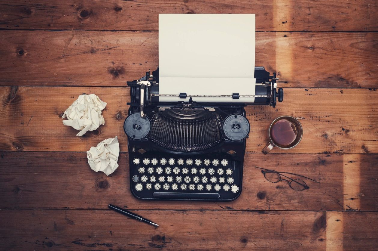 Old-fashioned typewriter on a wooden desk. Blank paper in typewriter. Two crumpled pages on desk. 
