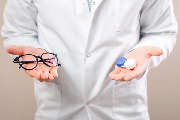 Person in white coat holding glasses in one hand and contact lens cases in the other.