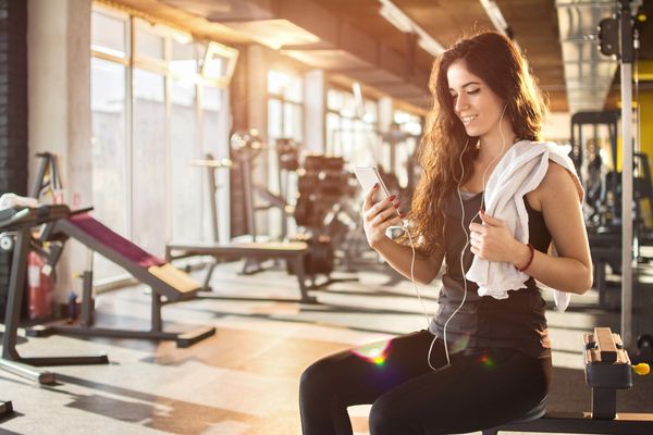 Young woman at gym checking phone with earphones and towel.