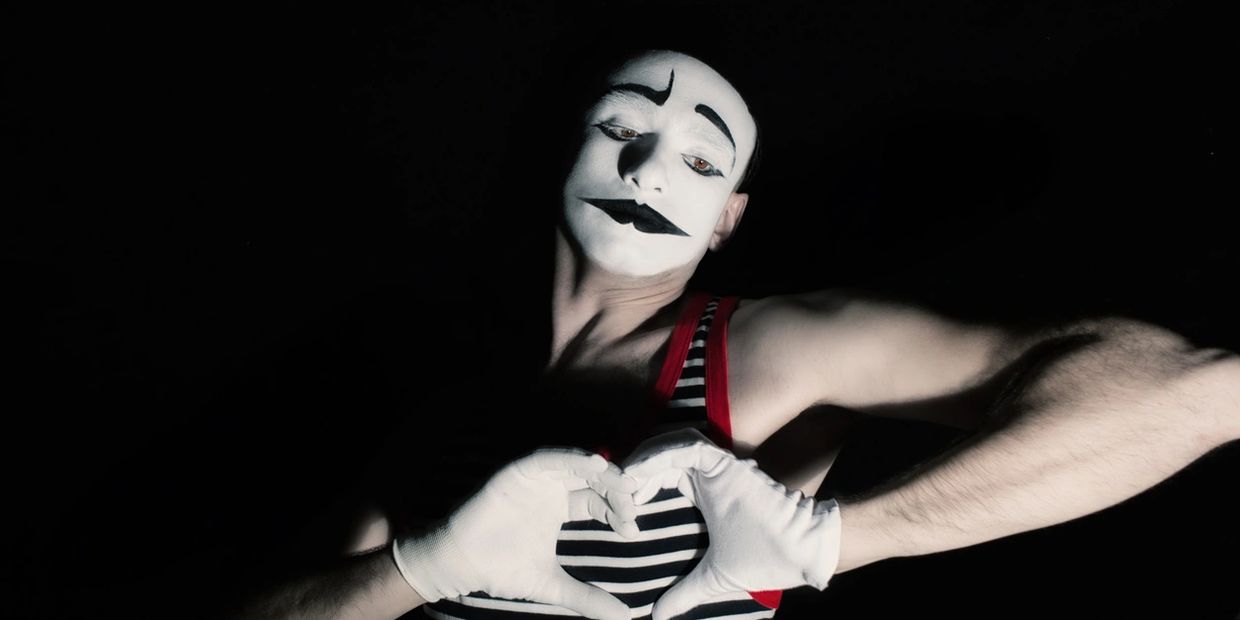 Mime artist making a heart shape with hands against a dark background.