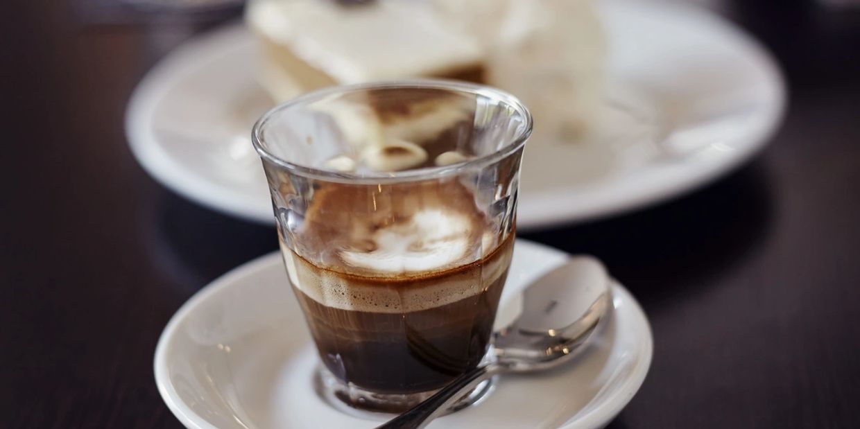 A glass of espresso with foam on a white saucer and spoon.