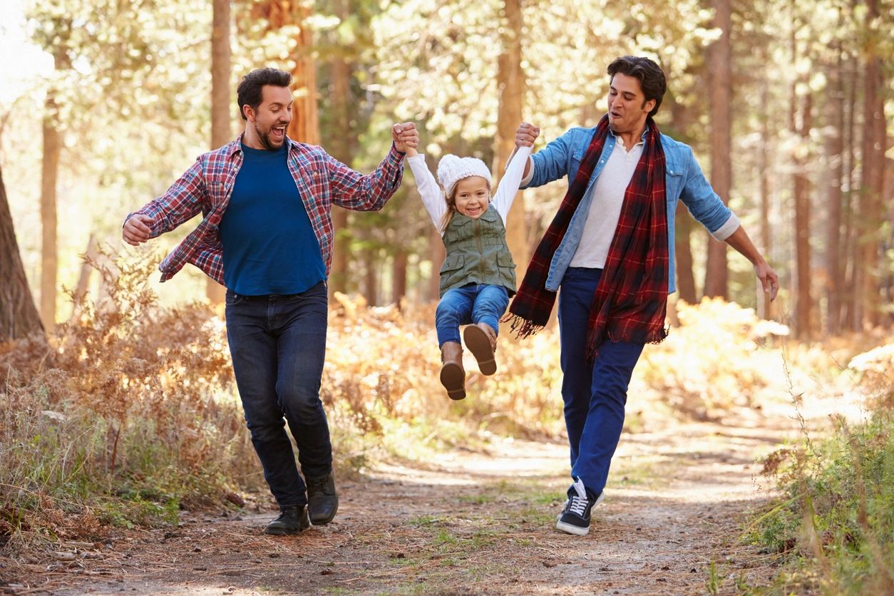 Two men joyfully swing a smiling child between them on a forest path.