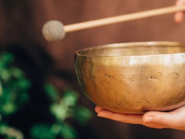 A hand holds a brass singing bowl with a mallet above it.