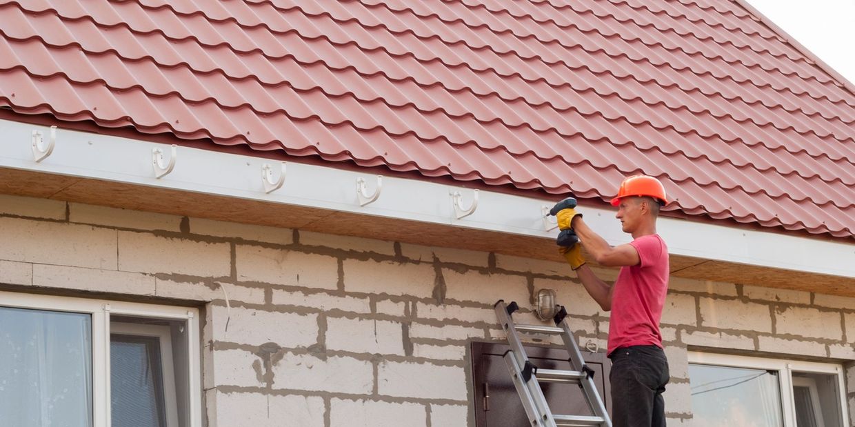 Man in a helmet fixing roof gutters using a drill on a ladder.