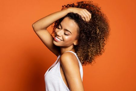 A joyful woman with curly hair poses against an orange background.