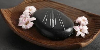 a wood tray containing a black rock with acupuncture needles on tops with orchids on each side