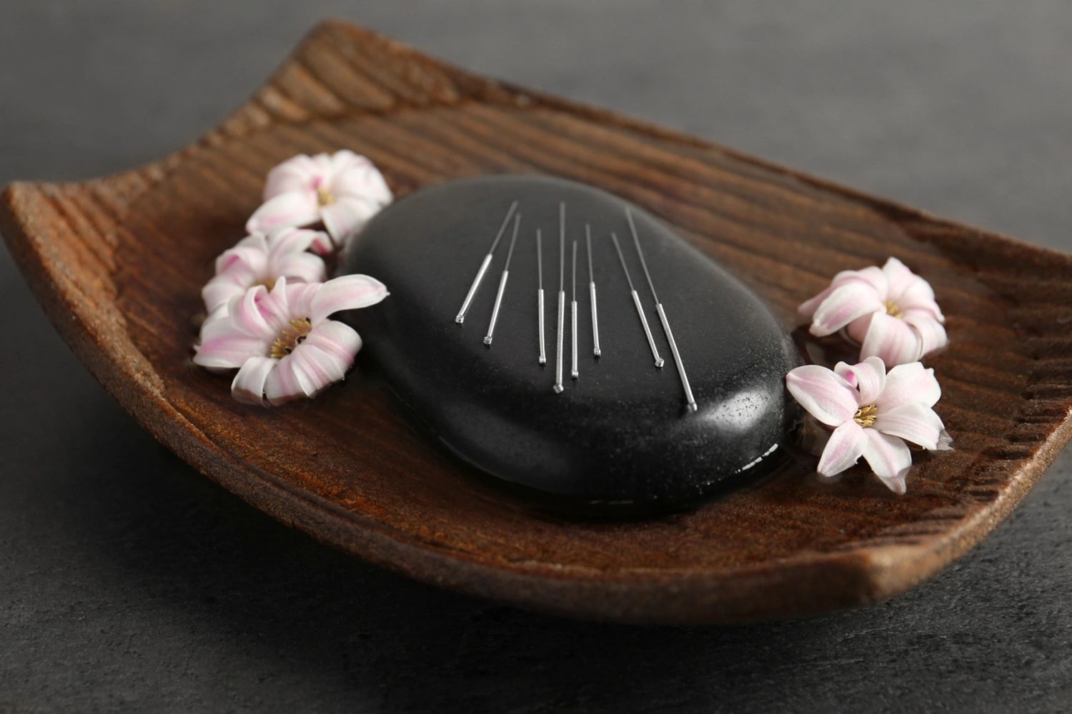 Stone with acupuncture needles on it and pink blossoms on a wood dish.