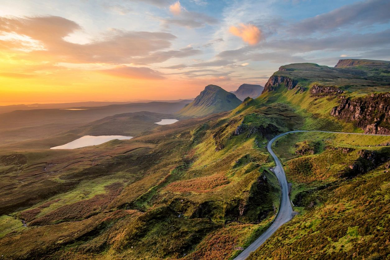 A winding road through green hills at sunset with lakes in the distance.