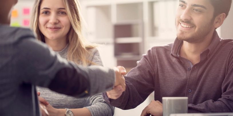 Two people shaking hands during a meeting while a woman watches and smiles.