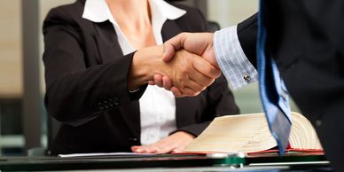 A seated woman in a suit shaking hands with a standing man in a suit and tie.