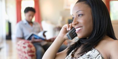 A woman on a phone call smiling with a man sitting in the background looking through a magazine.