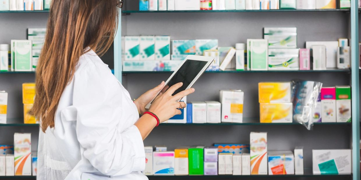 Pharmacist using a tablet in a pharmacy with medicine shelves in the background.