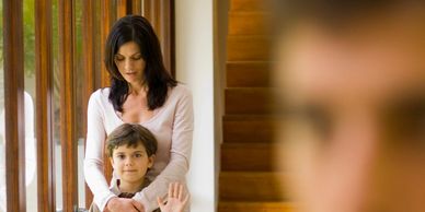 A mother stands behind her son, who waves at a man in the foreground.