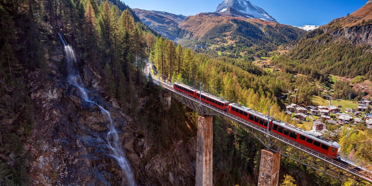 Red train crossing a bridge over a forested valley with a waterfall and mountain.