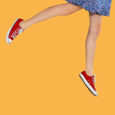 Person wearing red sneakers and a blue floral dress, jumping against an orange background.