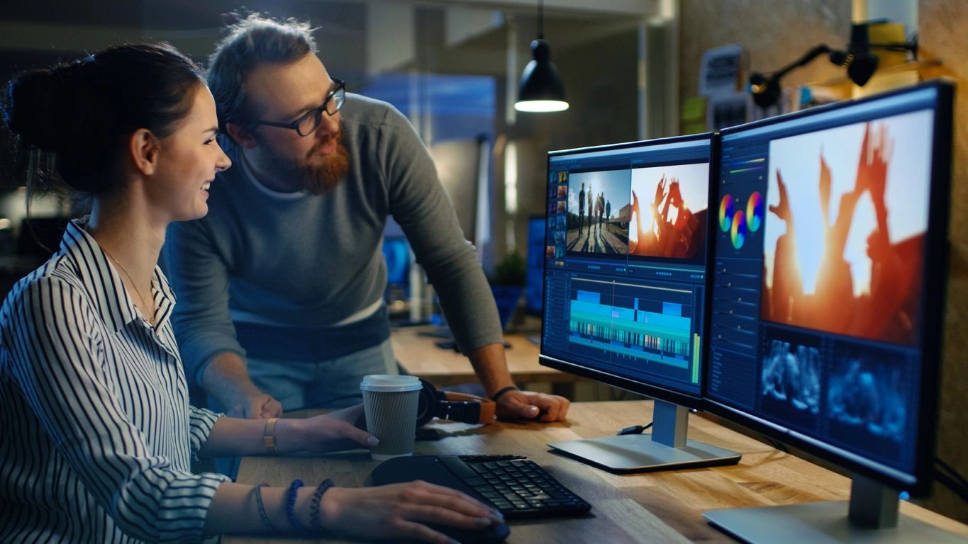 A woman and a man look at two computer screens at a desk.