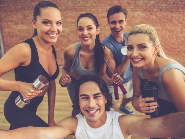 Group of five smiling people in workout clothes taking a selfie in a gym.