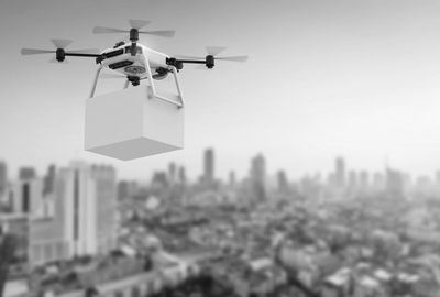 A drone carrying a package flies over a cityscape in black and white.