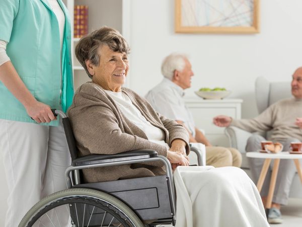 Elderly woman in wheelchair being assisted by a caregiver in a cozy living room.