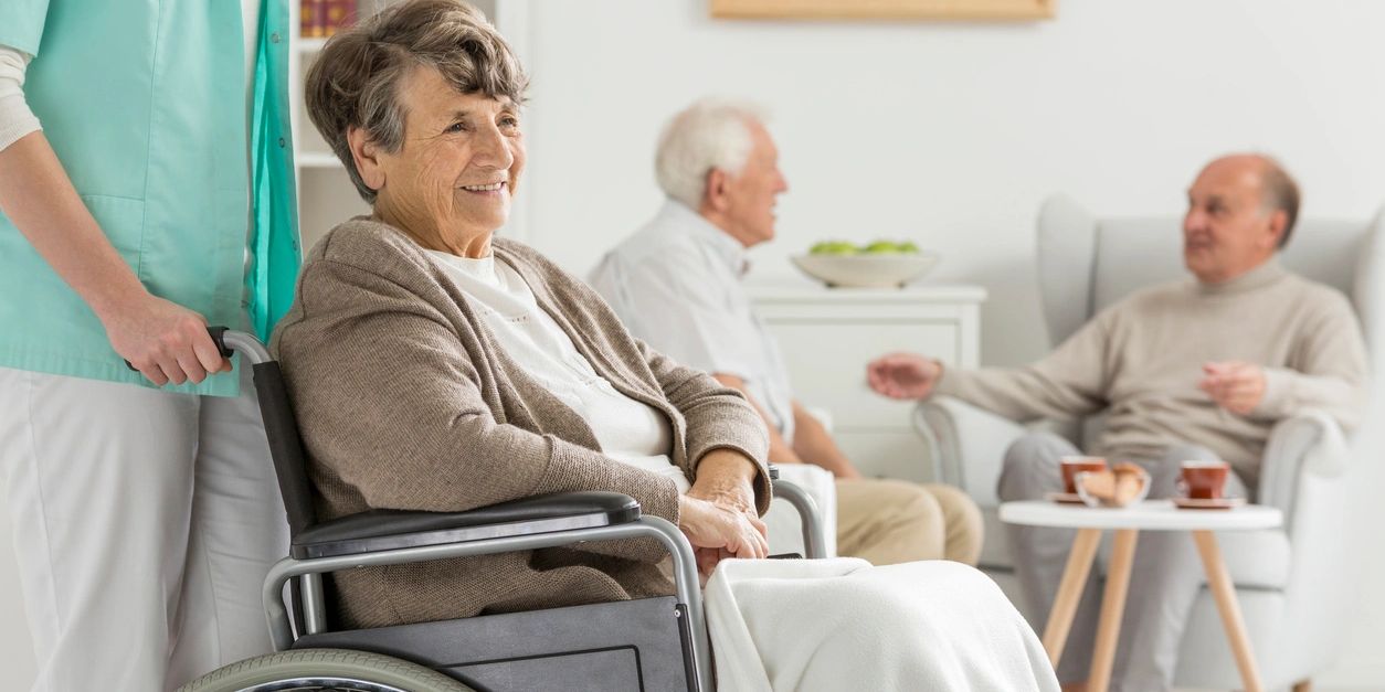 Elderly woman in a wheelchair beside a caregiver, two men chatting in the background