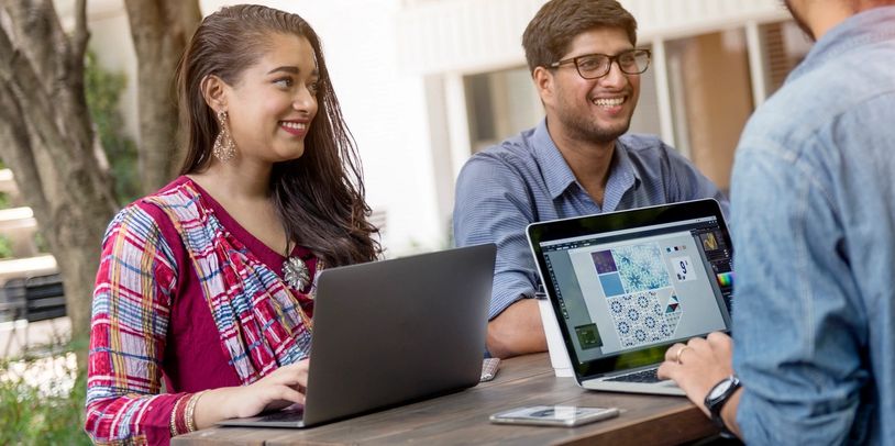 Three young professionals collaborating on laptops outdoors, smiling and engaged.