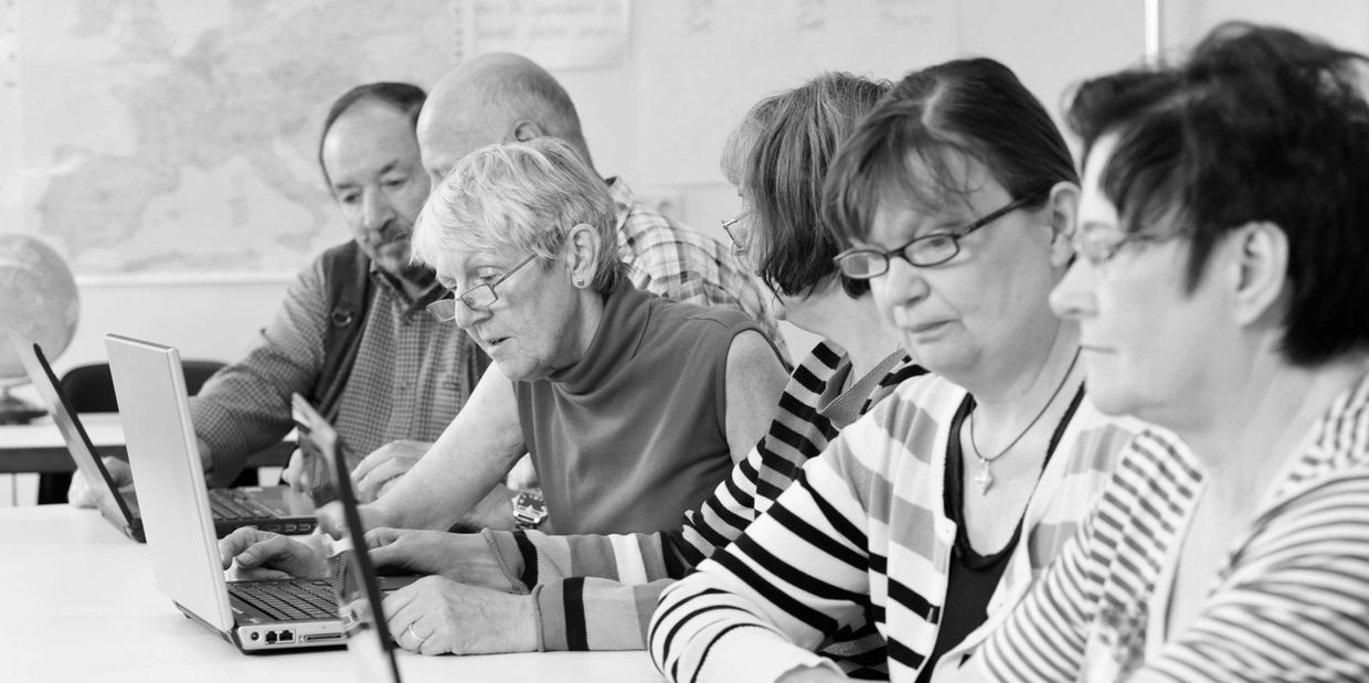 Group of mature people sharing laptops in a classroom style setting.