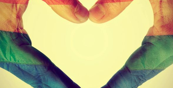 Hands forming a heart shape with a rainbow flag overlay symbolizing LGBTQ+ pride and love.