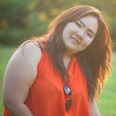 woman with long dark hair outside in front of trees, she's tilted to the side smiling at the camera