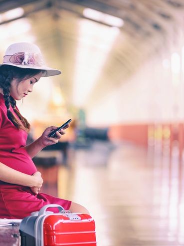 Pregnant woman in a red dress using phone at a station with red suitcase.