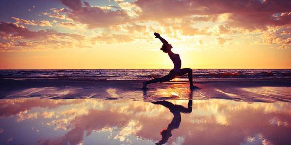 Woman in crescent lunge pose on sand and water surface with sunset and reflection.