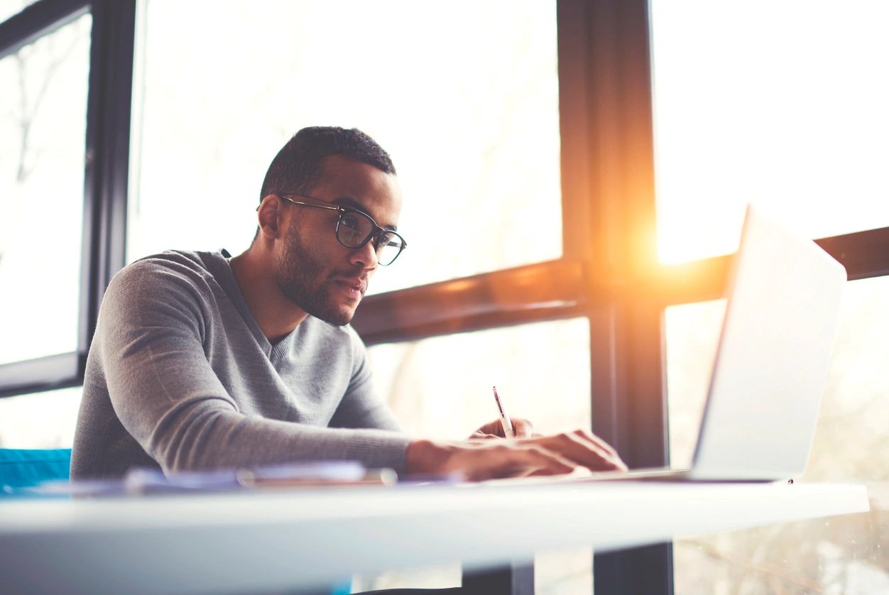 Young man with glasses on, focused on his study, as he searches his laptop while taking notes.