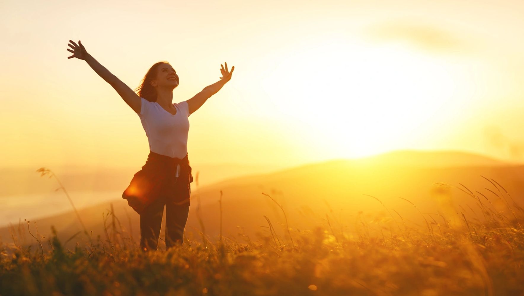 Woman joyfully raising arms in a sunlit field at sunset.