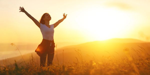 Woman standing in field on a hill at sunset with arms lifted up and outstretched in exuberance.