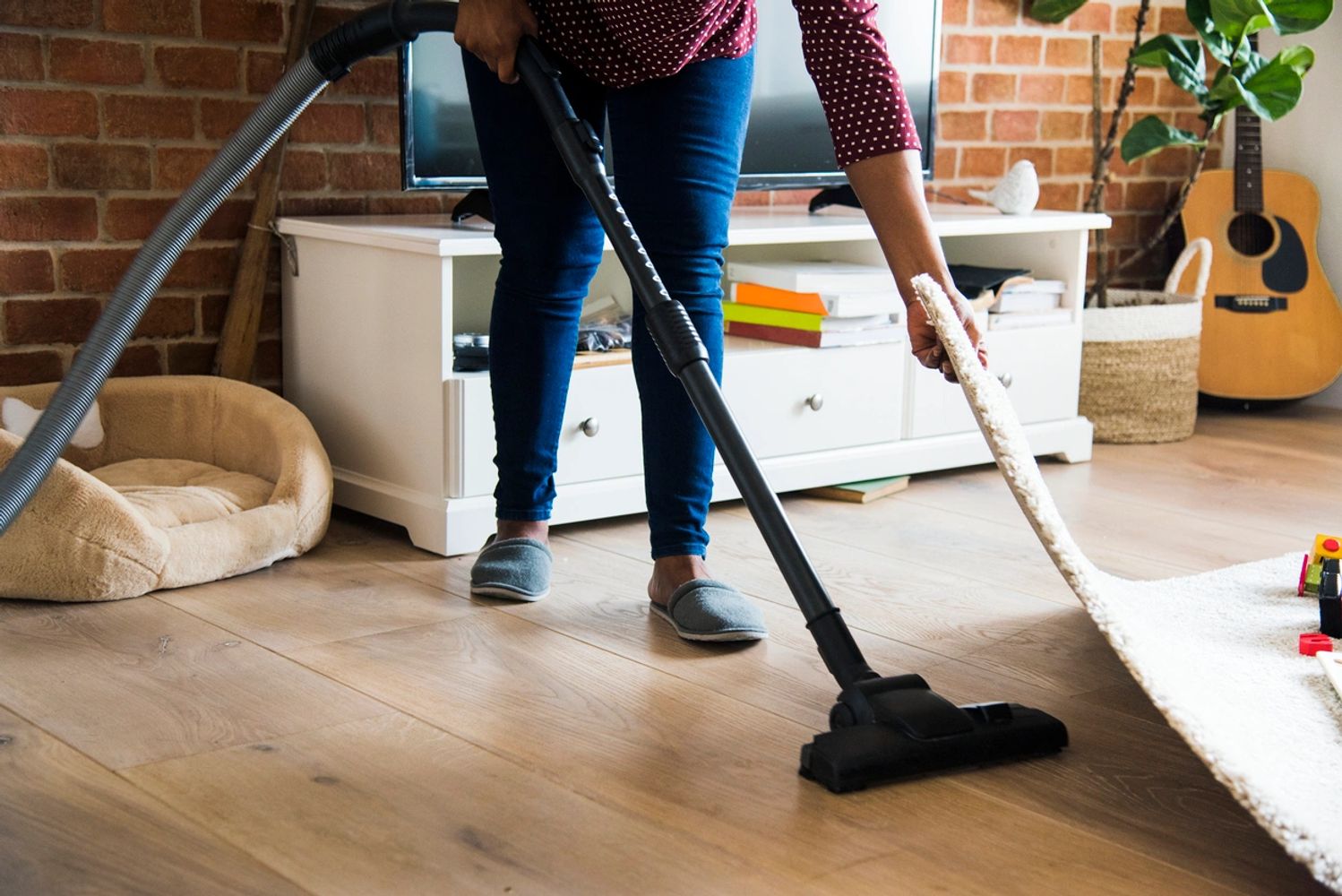 Person lifting carpet in order to vacuum under it, in a living room in front of a tv and a pet bed.