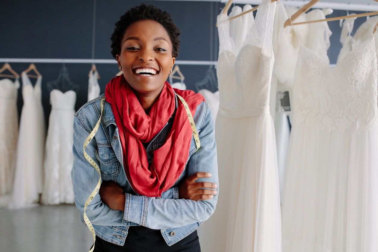 A seamstress smiles with her tape measure over her shoulders and bridal gowns behind her.