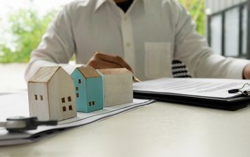 Miniature house models on documents with a person in the background.