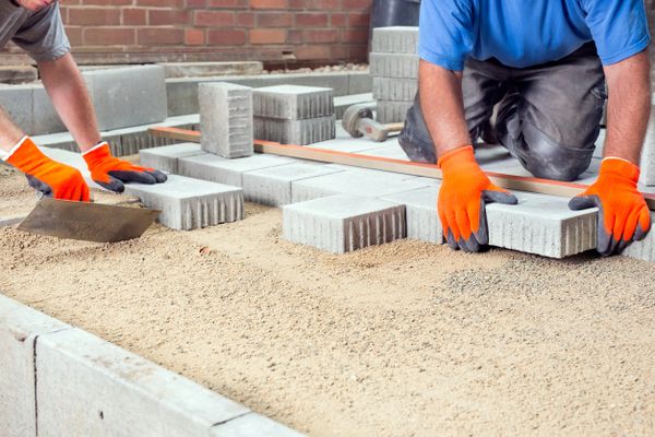 Workers laying concrete pavers on sand with orange gloves.