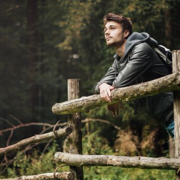 Young man in a leather jacket leaning on a wooden fence in a forest.