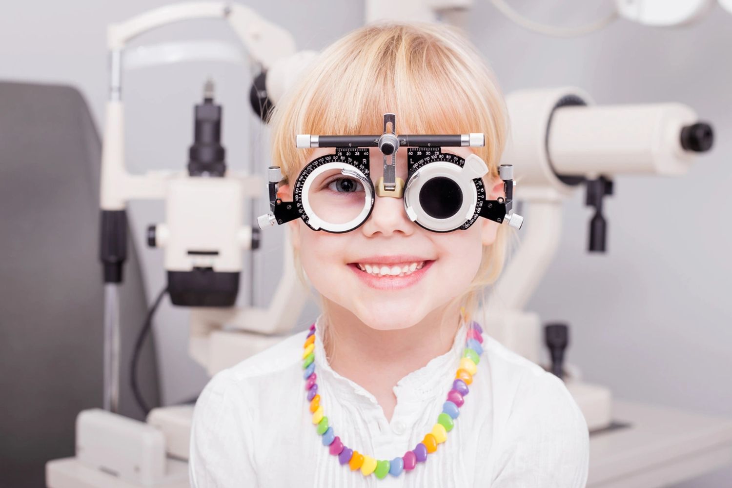 Young girl wearing a white shirt with a rainbow necklace and an optometrist trial frame, smiling. 