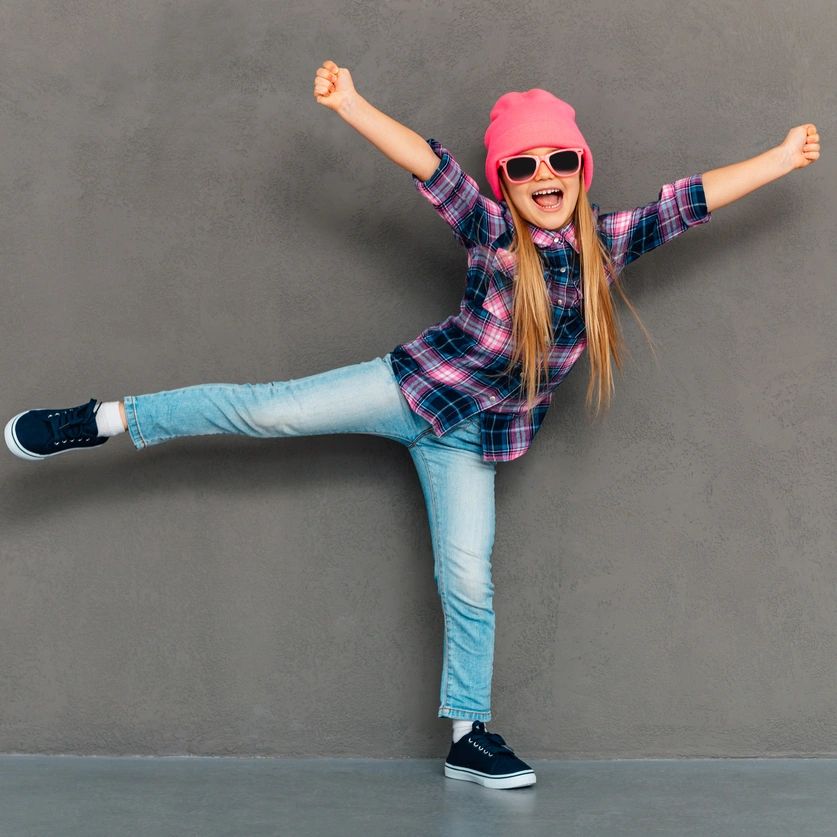 Joyful girl in pink hat and sunglasses striking a playful pose against a gray wall.
