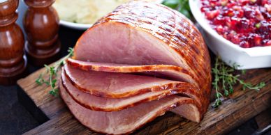 Sliced glazed ham on a wooden board with cranberry sauce and seasonings.