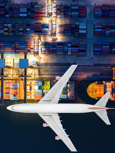 Aerial view of a large airplane flying over a brightly lit cargo port with containers.