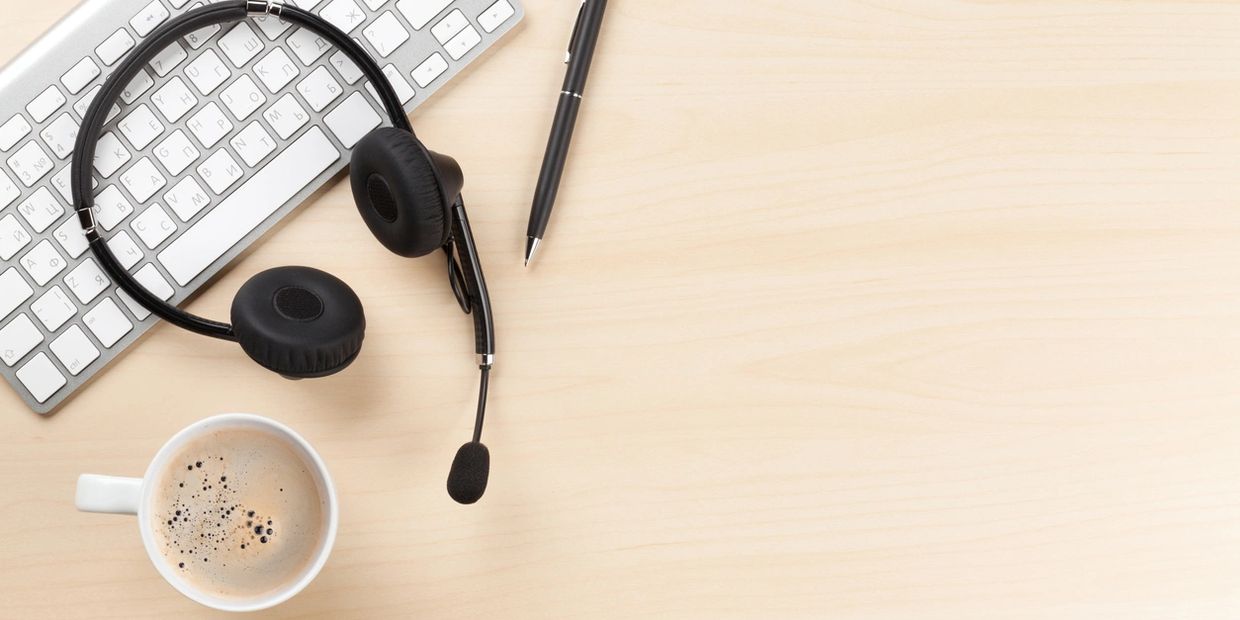 Workspace with headset, keyboard, pen, and coffee on a light wooden desk.