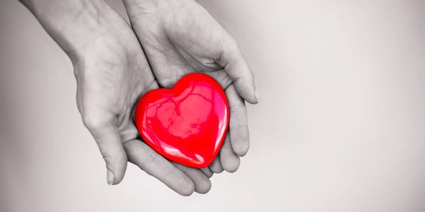 Black and white image of two hands holding a red ceramic heart.