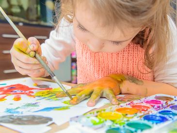Girl painting the back of her hand with a paint brush and bright colours wearing an apron at a table